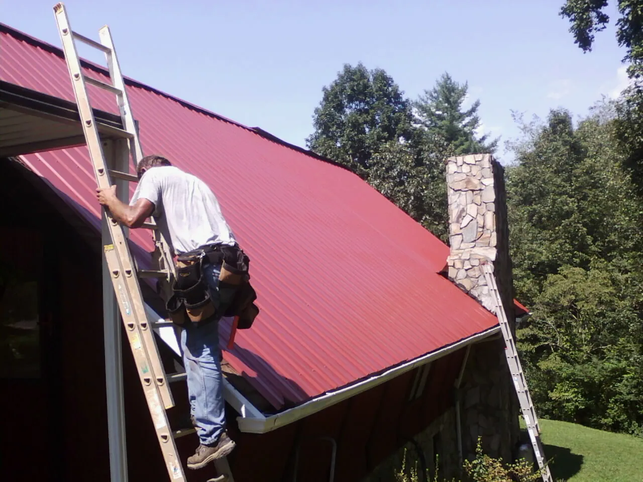 Roofer on a ladder beside a red metal roof and stone chimney — Soffit & Fascia Repair by Roofing Brothers in Royalton